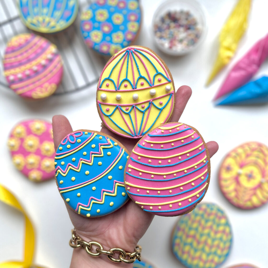 Pretty iced Easter Egg biscuits in someone's hand, with icing bags and decorations in the background, from the Easter Egg DIY icing kit by Baked by Steph