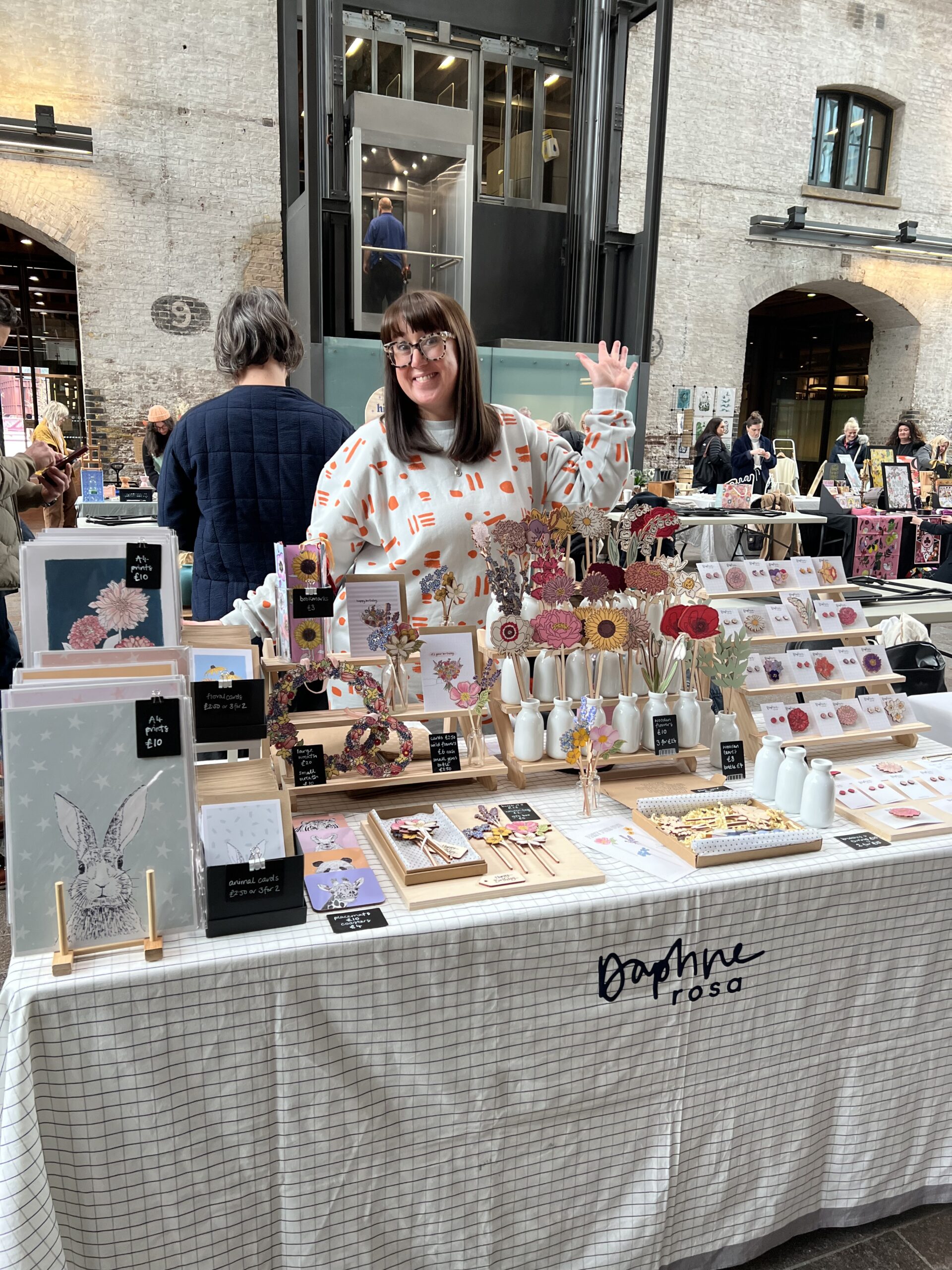 Kate from DaphneRosa at a market, standing behind her stall with prints, wooden flowers, and homewares on display.