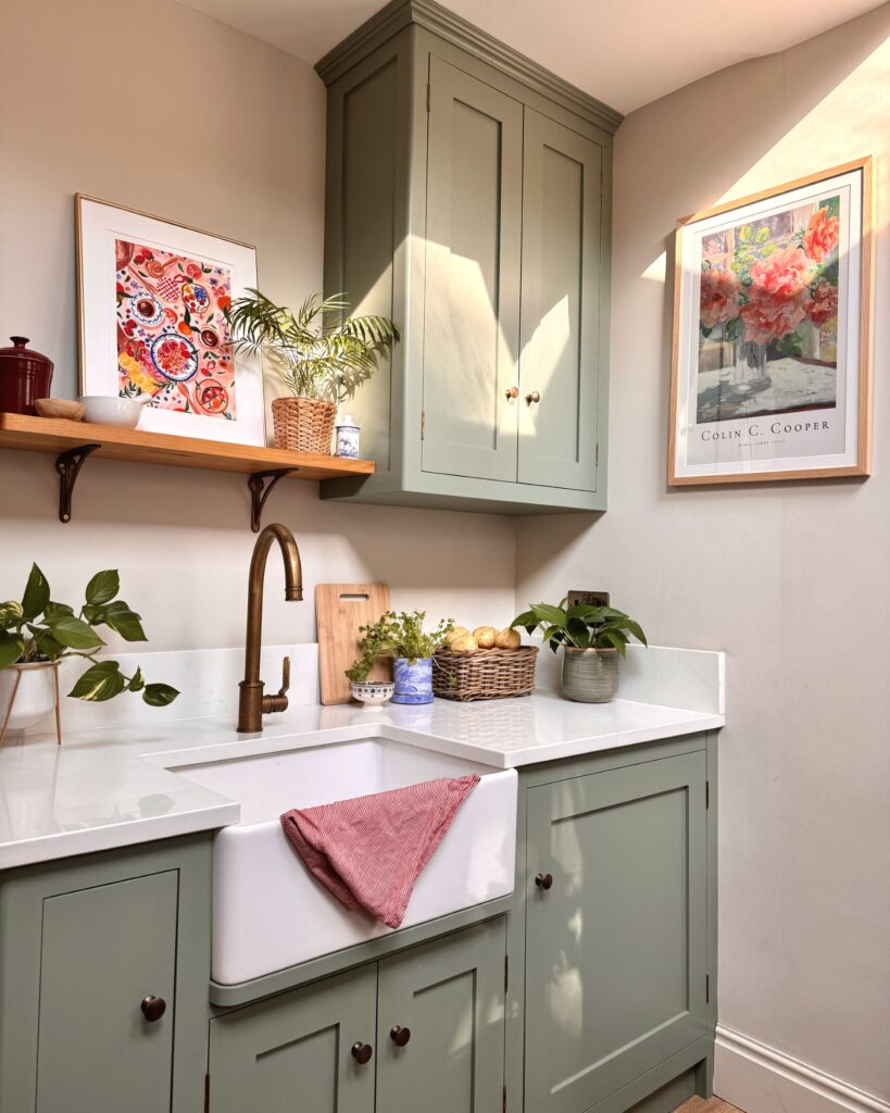 A kitchen sink area designed by Becks from The Interior Lens, with soft muted green cupboards, a large sink, and touches of colour from plants to artwork