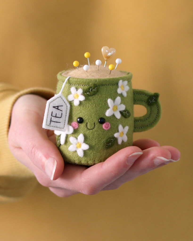 Green mug pincushion with white daisies, a little smiley face, and a 'tea' tag, in the hand of its creator: hannahdoodle