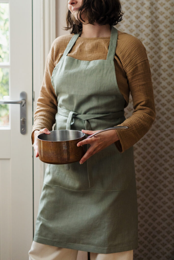 Model wearing a sage green linen apron by Charlotte from Cloth & Clay, holding a saucepan