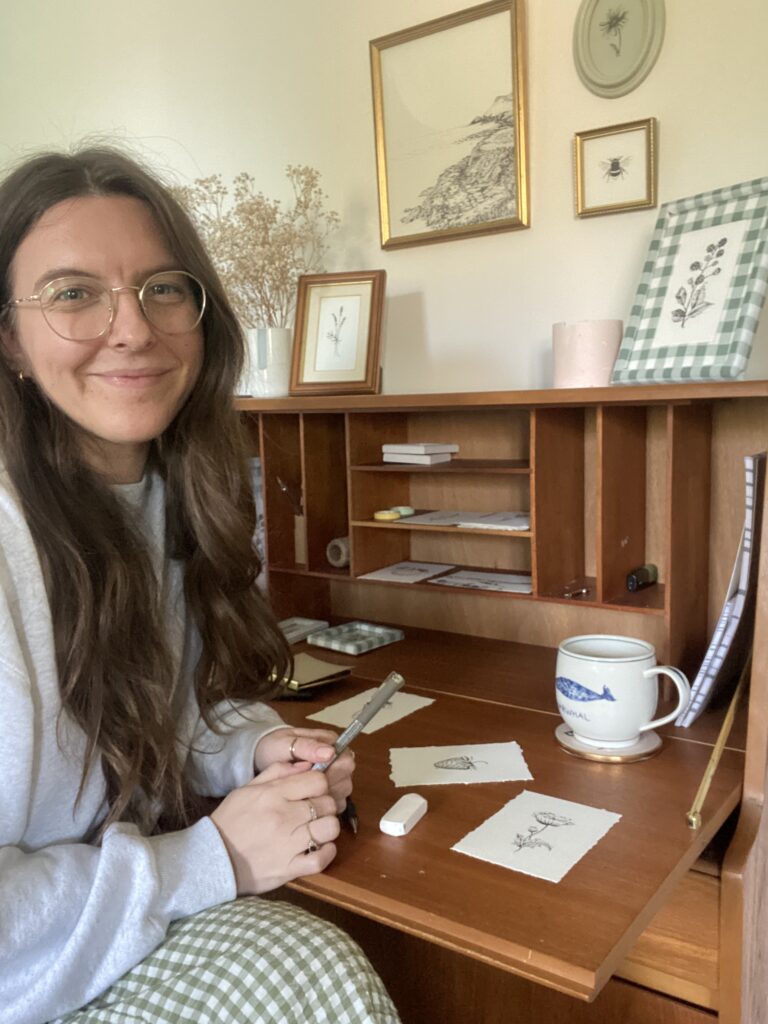 Catherine from Catherine Bamber Art sitting at her desk with pieces of art and a cup of tea in front of her