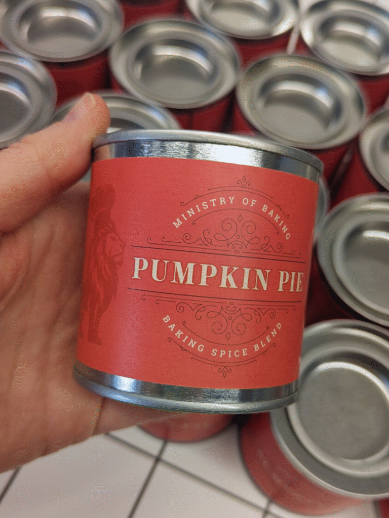 A hand holding a tin of Pumpkin Pie Baking Spice Blend by Ministry of Baking, with more tins in the background