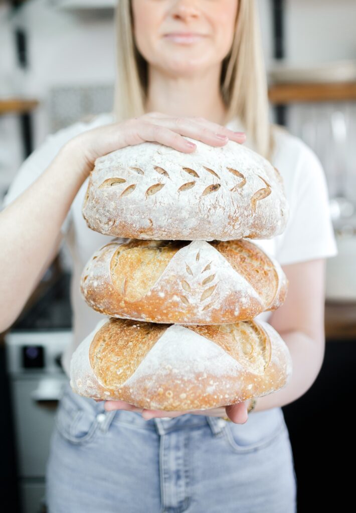 Charlotte from British Sourdough holding three sourdough loafs on top of each other, each with a decorative pattern scored into the surface.