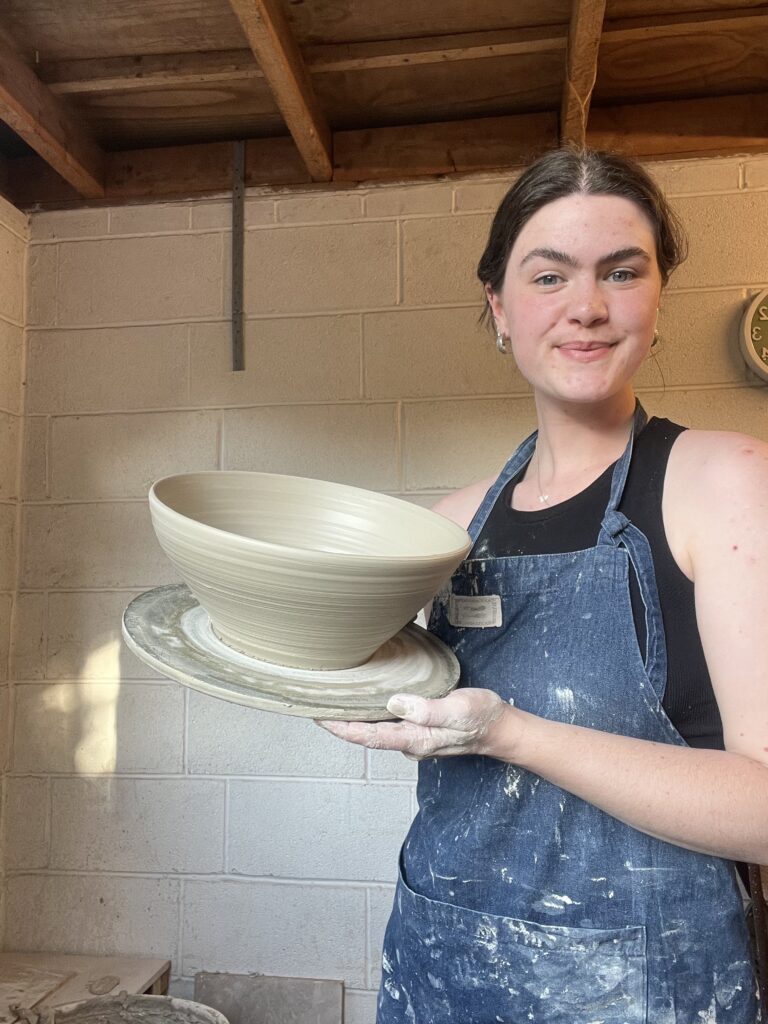 Kat from Ceramics by Kat, wearing an apron, holding one of her wheel-thrown potter pieces