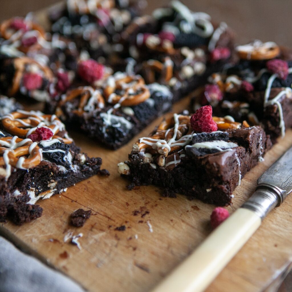 Brownie slices loaded with toppings, on a wooden chopping board with a knife by the side, by Honeywell Bakes