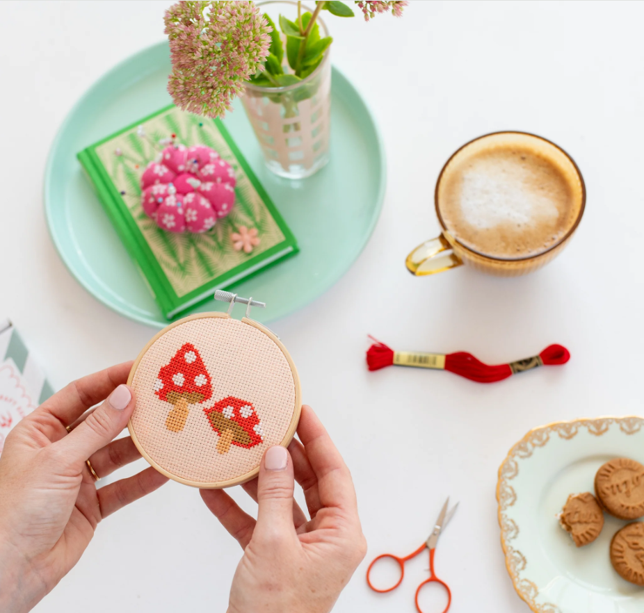 Red toadstool cross stitch in hoop, by Cotton Clara, being held by two hands, with flowers, coffee, thread, scissors, and biscuits on the table behind