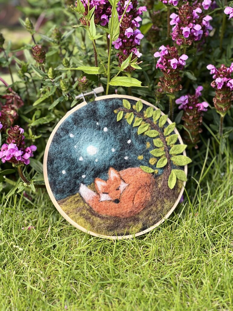 Sleepy Fox needle felting painting with wool, in a hoop, placed in a garden with grass and flowers around