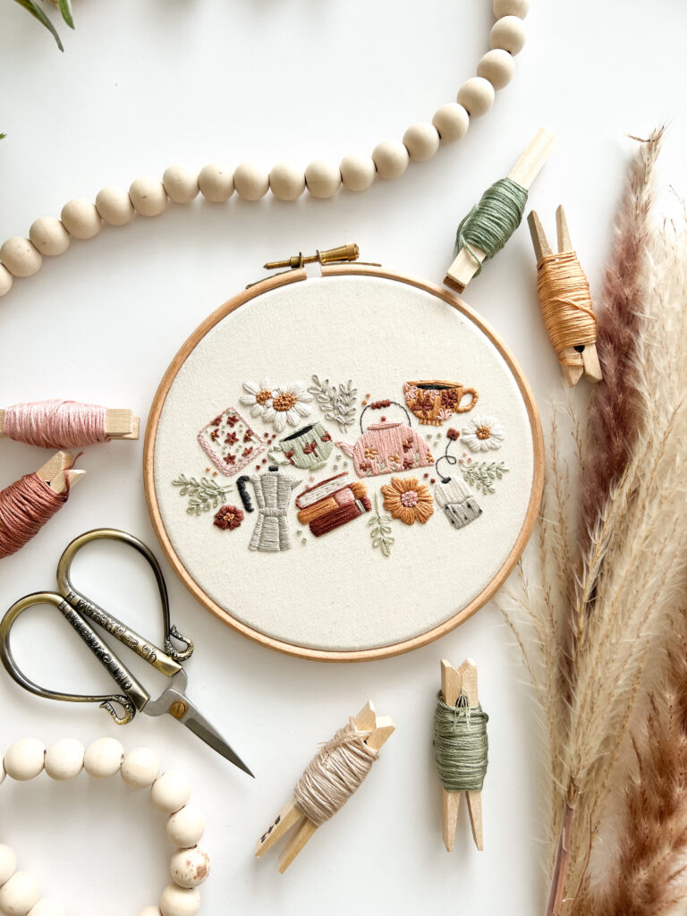 Tea coffee books and flowers embroidery pattern by Mindful Mantra Embroidery - stitched in warm neutral tones, in a hoop, surrounded by antique scissors, threads, and neutral props