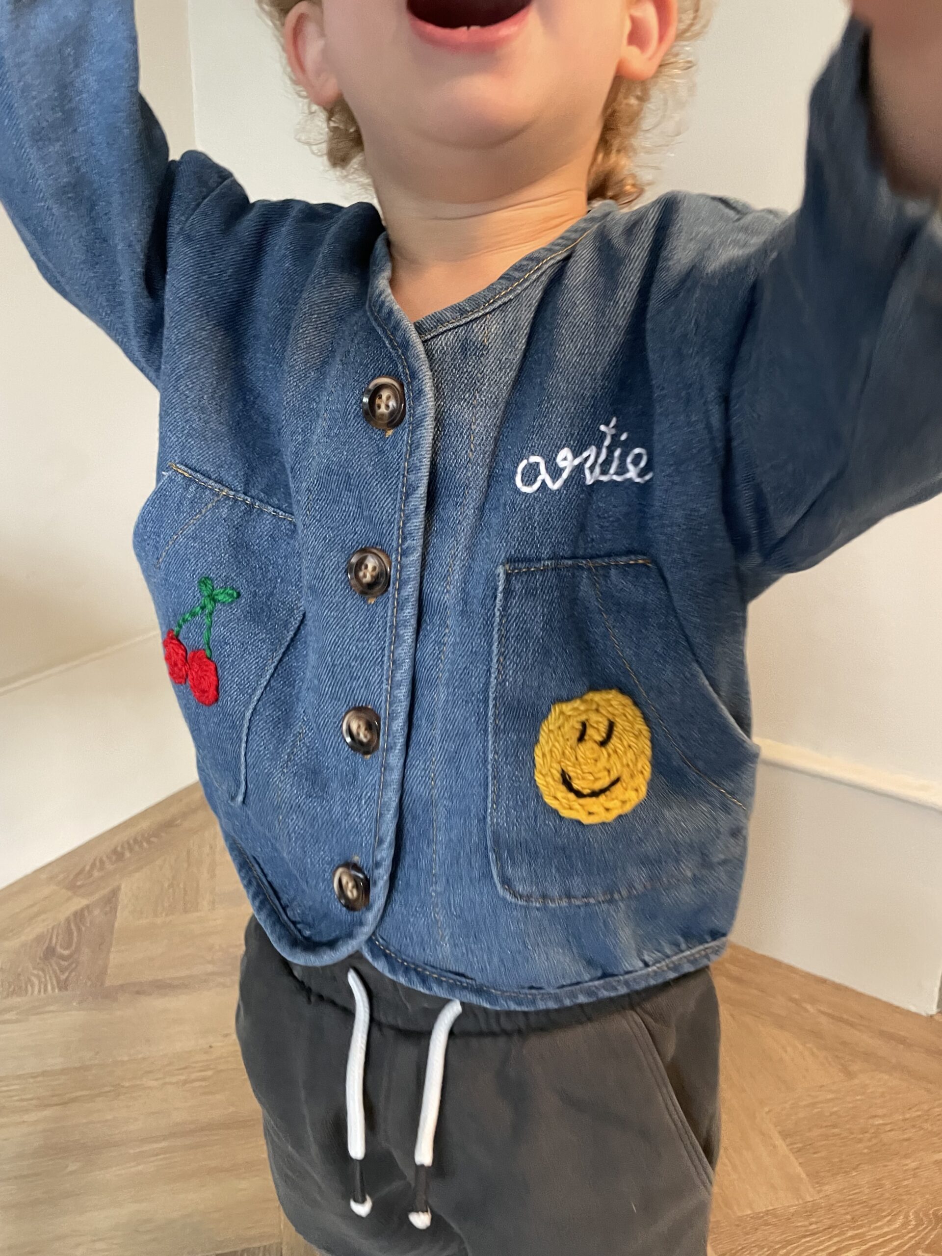 One of Jay's embroidered kidswear creations from By Jay - a denim cardigan jacket with an embroidered smiley face, cherries, and name