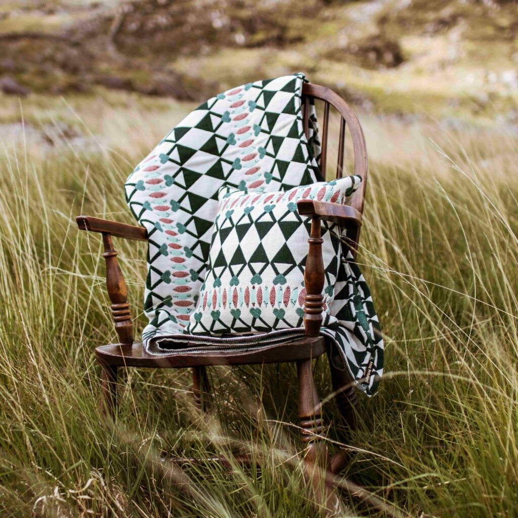 A blanket and cushion cover by Anna Pritchard, a textile designer in North Wales, on a wooden chair in a gassy field