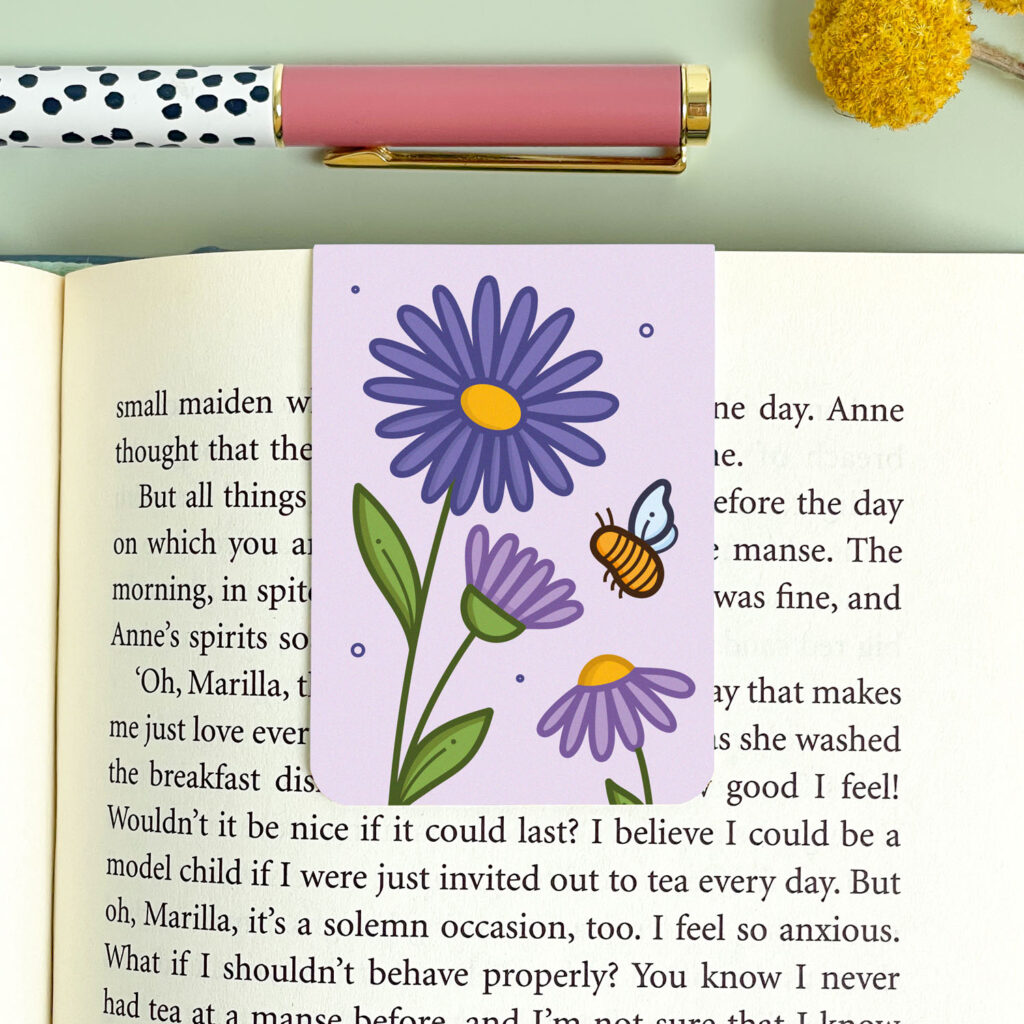 A September birth flower magnetic bookmark with a purple flower and a bee design, folded over a book page. Created by Joanne Hawker.