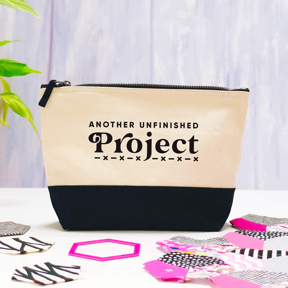 A project bag pouch by Joanne Hawker with the words 'Another unfinished project' on the front, with fabric pieces on the table next to it.
