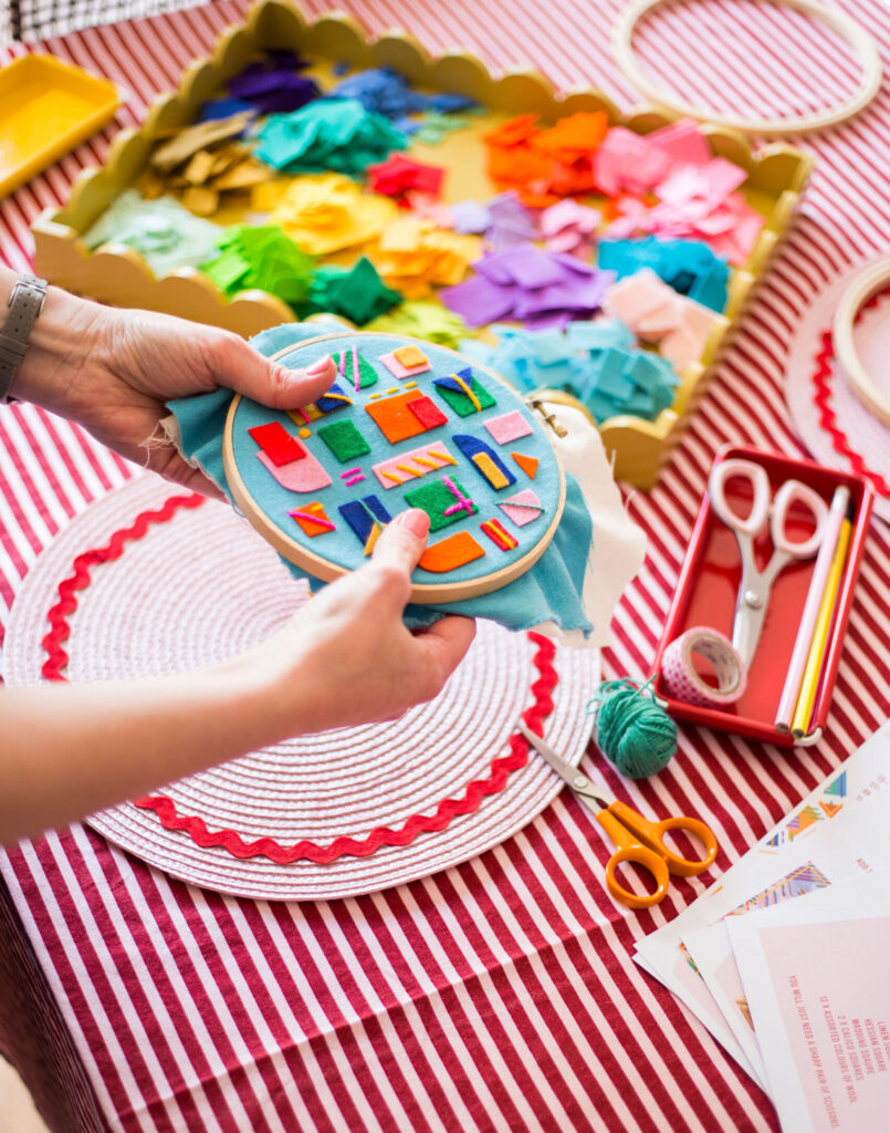 Abstract embroidery kit by Miss Shelly Designs with a pair of hands holding an embroidery hoop with colourful felt and stitching, above a table with a red and white striped table cloth