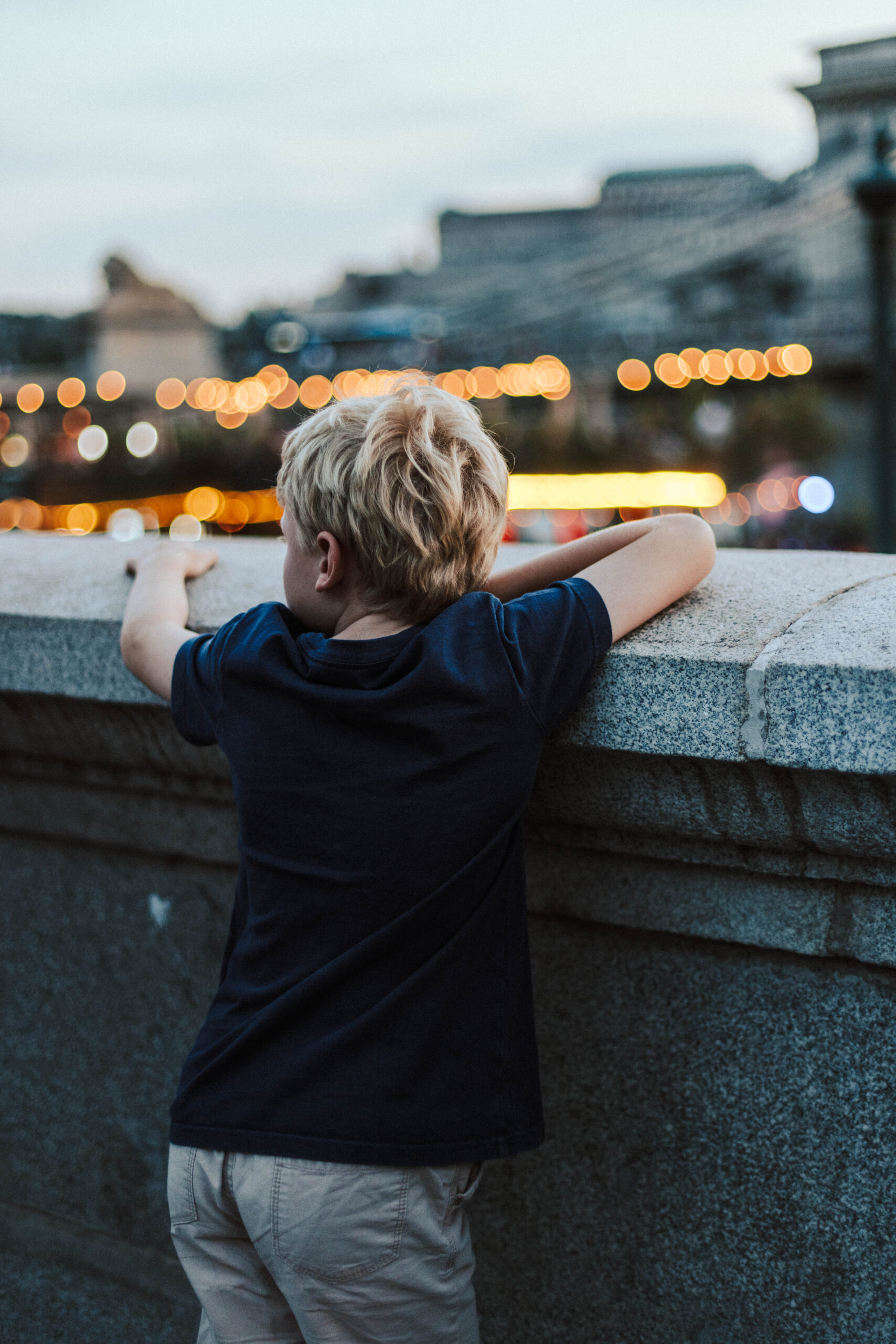 A photo by York family and brand photographer, Sarah Burton Photography, of a young boy leaning on a brick wall, looking across a river towards a cityscape (with blurred buildings and lights in the distance).