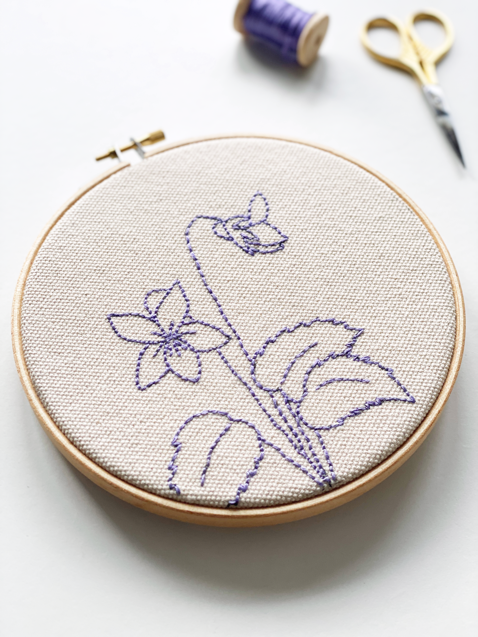 February birth flower, violet embroidery finished project in a wooden hoop, on natural cotton canvas with purple embroidery thread, with gold embroidery scissors and a wooden bobbin of thread in the background, by For Creatives' Sake