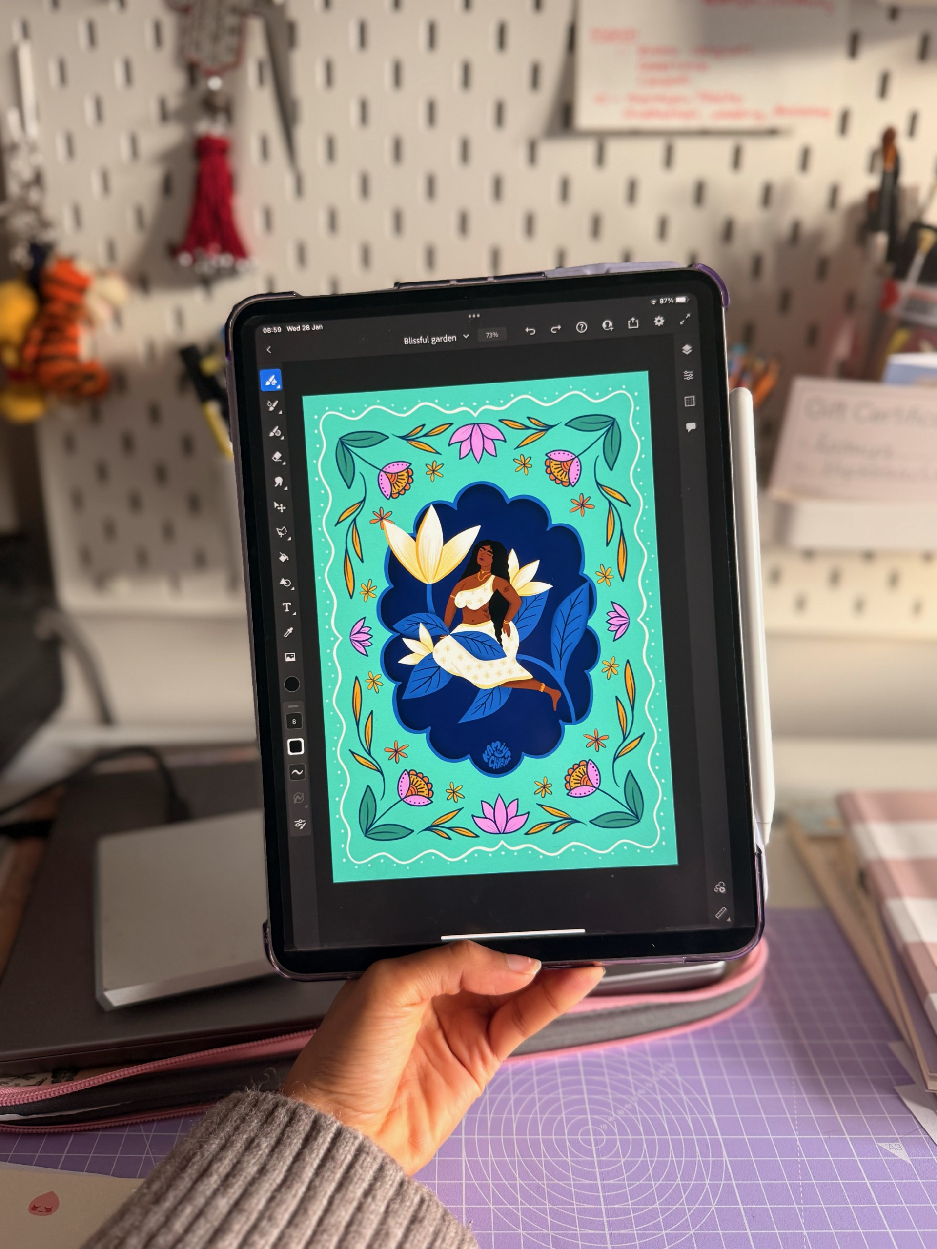 A woman holding an iPad with a Kamiya Chirodian illustration on the screen (of a South Asian woman surrounded by florals, with a turquoise, blue, pink, and cream colour palette)