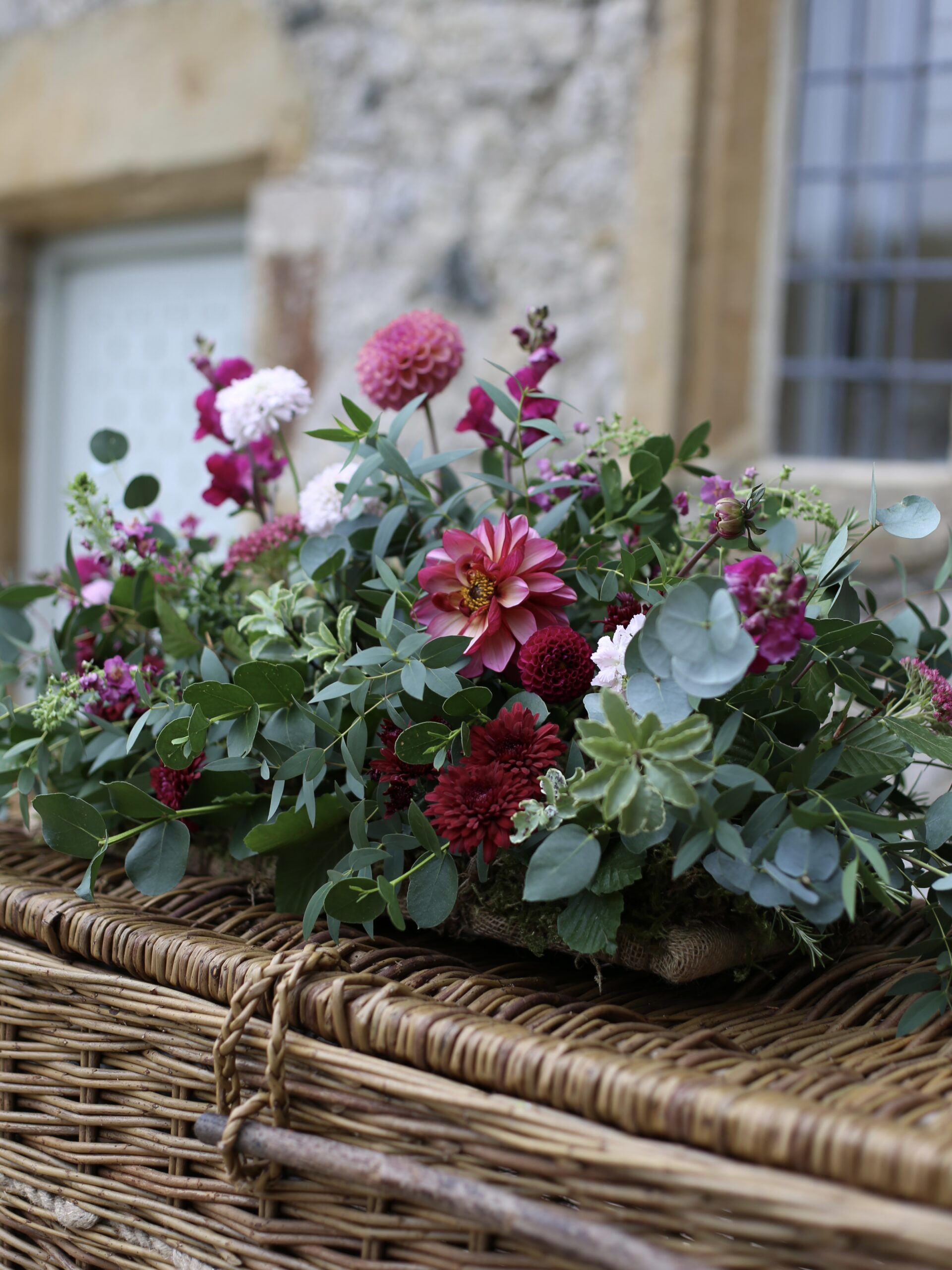 A funeral arrangement on a coffin, with dark red and pink flowers and lots of greenery, by Tracey from Wild Soul Flowers