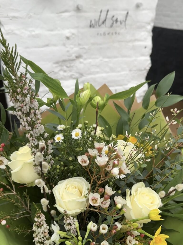 A bouquet by Wild Soul Flowers of white and soft pink and yellow flowers, plus greenery, with the logo in the background