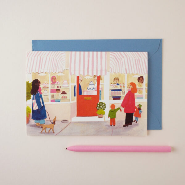 Patisserie bakery illustrated card by Peggy & Kate, with illustration (originally hand illustrated in gouache and pencil) of a small patisserie bakery with a red door, striped awning, and women passing by. A pink pen is next to the card. The envelope is mid-light blue.