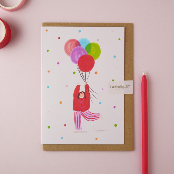 Birthday Balloons illustrated card by Martha Ratcliff Illustration, with an illustration of a woman holding balloons floating into the air on the front. A red pen and red and pink washi tapes are next to the card. The envelope is brown kraft.