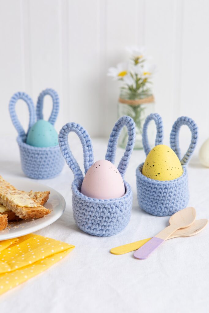 Three light blue crocheted bunny-ear egg cups by Wool Couture, with a pastel coloured egg in each, and a mock breakfast table scene with toast on a plate, spoons, and flowers in the background