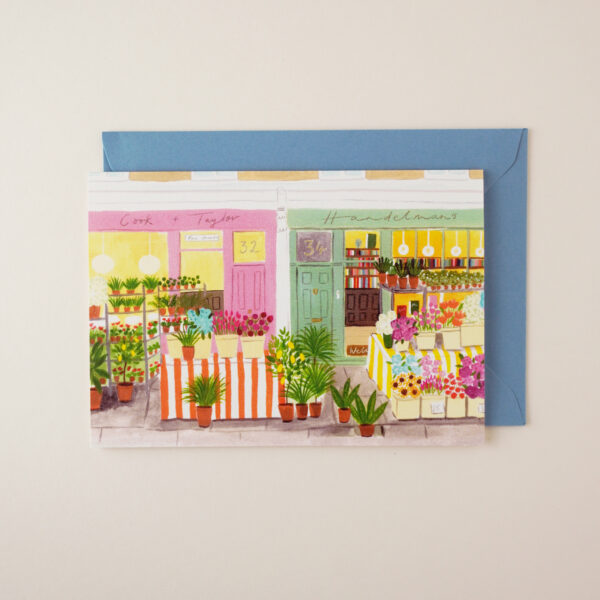 Flower market shop illustrated card by Peggy & Kate, with illustration (originally hand illustrated in gouache and pencil) of a small florist stall outside shops with pink and green doors. The envelope is mid-light blue.
