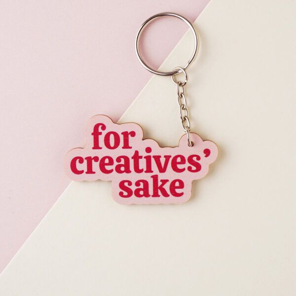 A wooden keyring, shaped around the brand name For Creatives' Sake, with a pink background and red text, with a silver keychain.