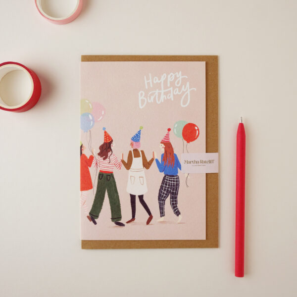 Happy Birthday friends card by Martha Ratcliff Illustration, with an illustration of four women partying wearing party hats on the front. A red pen and red and pink washi tapes are next to the card. The envelope is brown kraft.