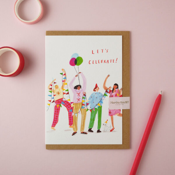 "Let's Celebrate" card by Martha Ratcliff Illustration, with an illustration of four women partying on the front. A red pen and red and pink washi tapes are next to the card. The envelope is brown kraft.