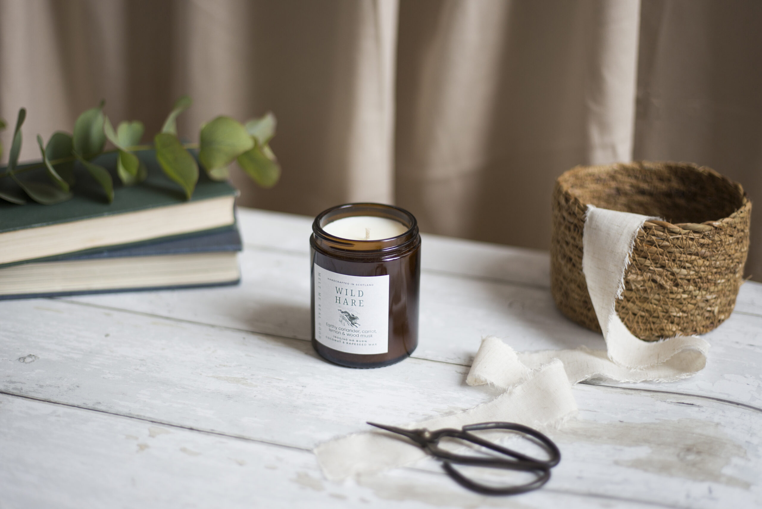 A brand product photograph by Lucy Hannah Photography of a candle in a brown glass container, with a pair of traditional scissors on the table, a ribbon, books, and some greenery