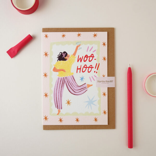 "Woo Hoo" card by Martha Ratcliff Illustration, with an illustration of a woman leaping through the air with her arm up on the front. A red pen and red and pink washi tapes are next to the card. The envelope is brown kraft.