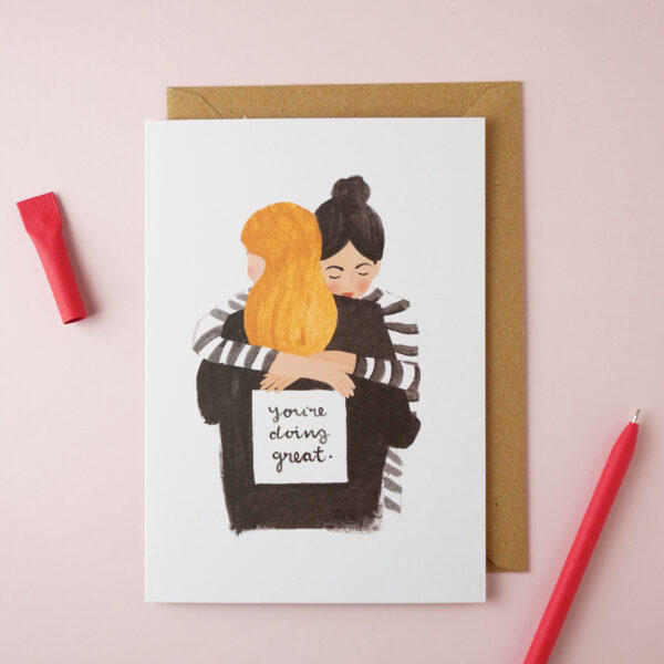 "You're doing great" card by Jade Fisher, with an illustration of two women friends hugging on the front. A red pen is next to the card. The envelope is brown kraft.